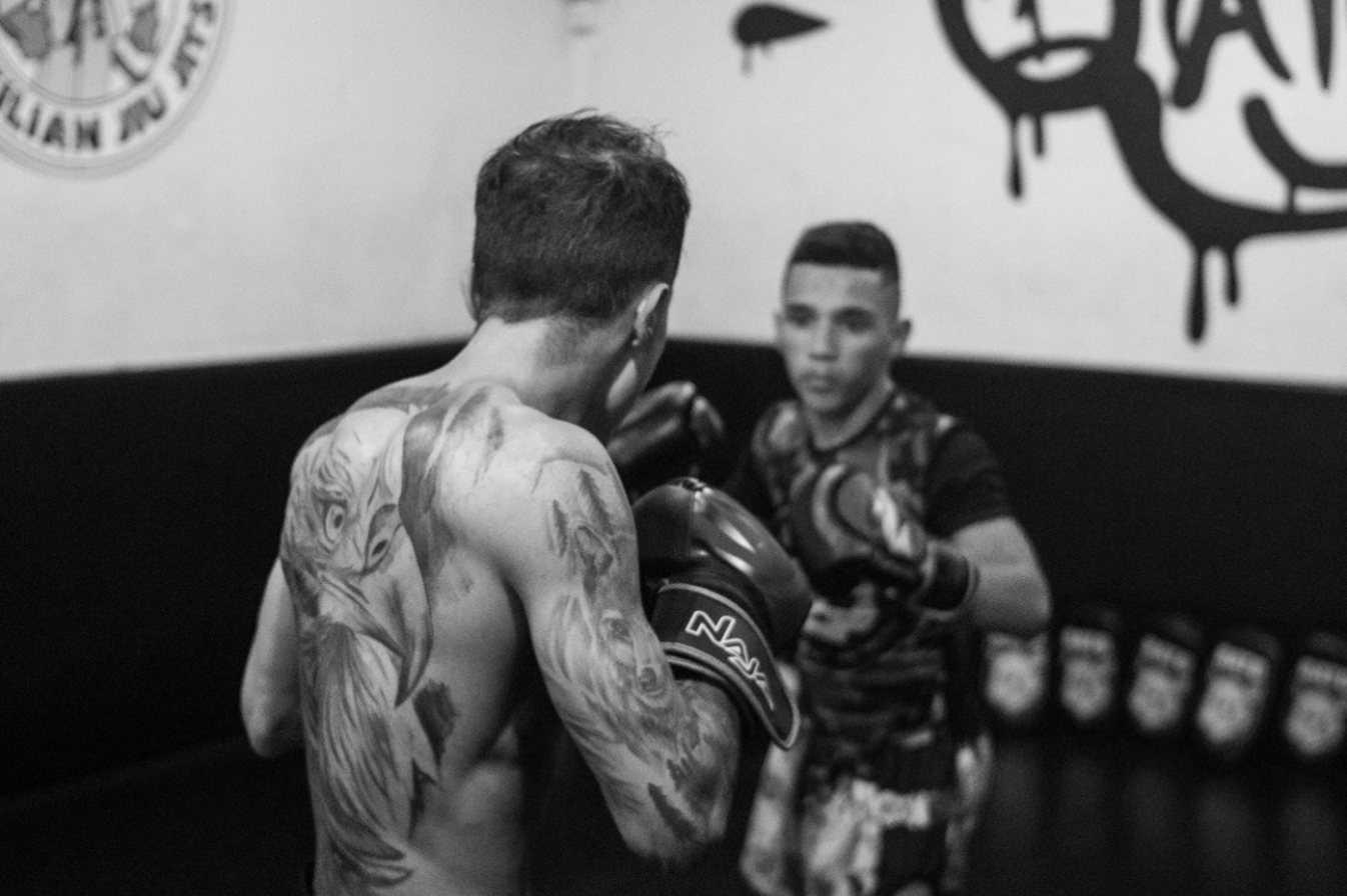 Dois lutadores em treino, um deles de costas com o braço tatuado, em preto e branco