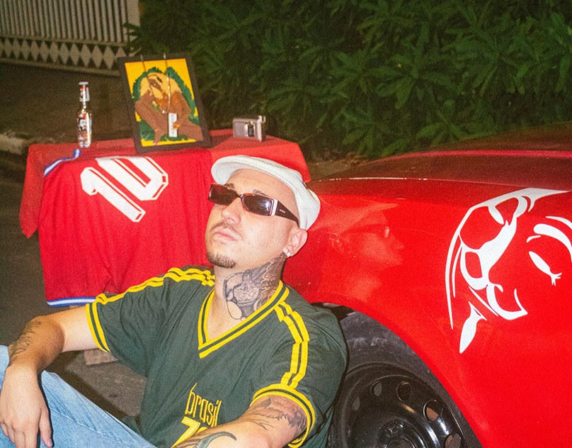 Homem de óculos escuros e boné branco encostado em um carro vermelho, ao lado de um altar com camisa de futebol